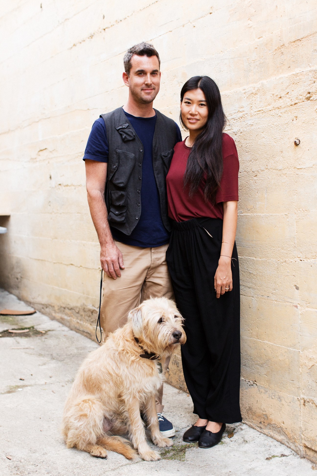 Two people stand with a cream-colored dog against a beige brick wall in an alley.
