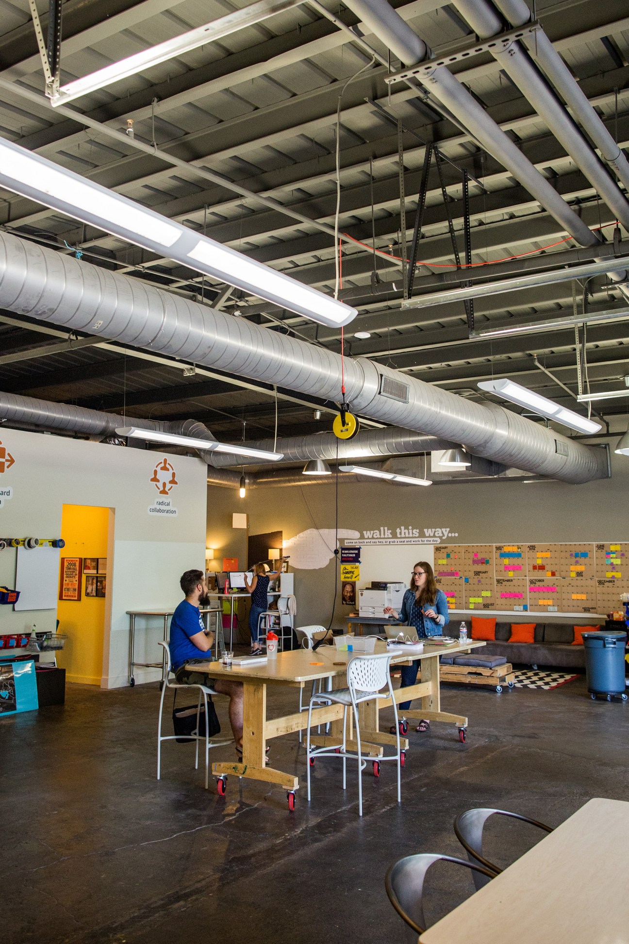 Interior of The Skillery co-working space with exposed industrial ceiling, wooden tables, and workers collaborating.