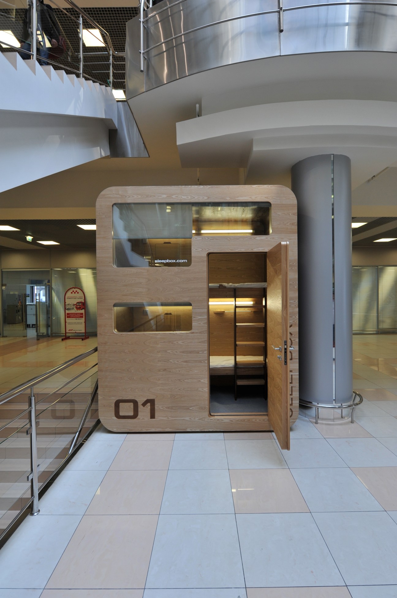 Sleepbox mini-hotel pod with ash wood exterior and open door displaying interior bunk bed at airport terminal.