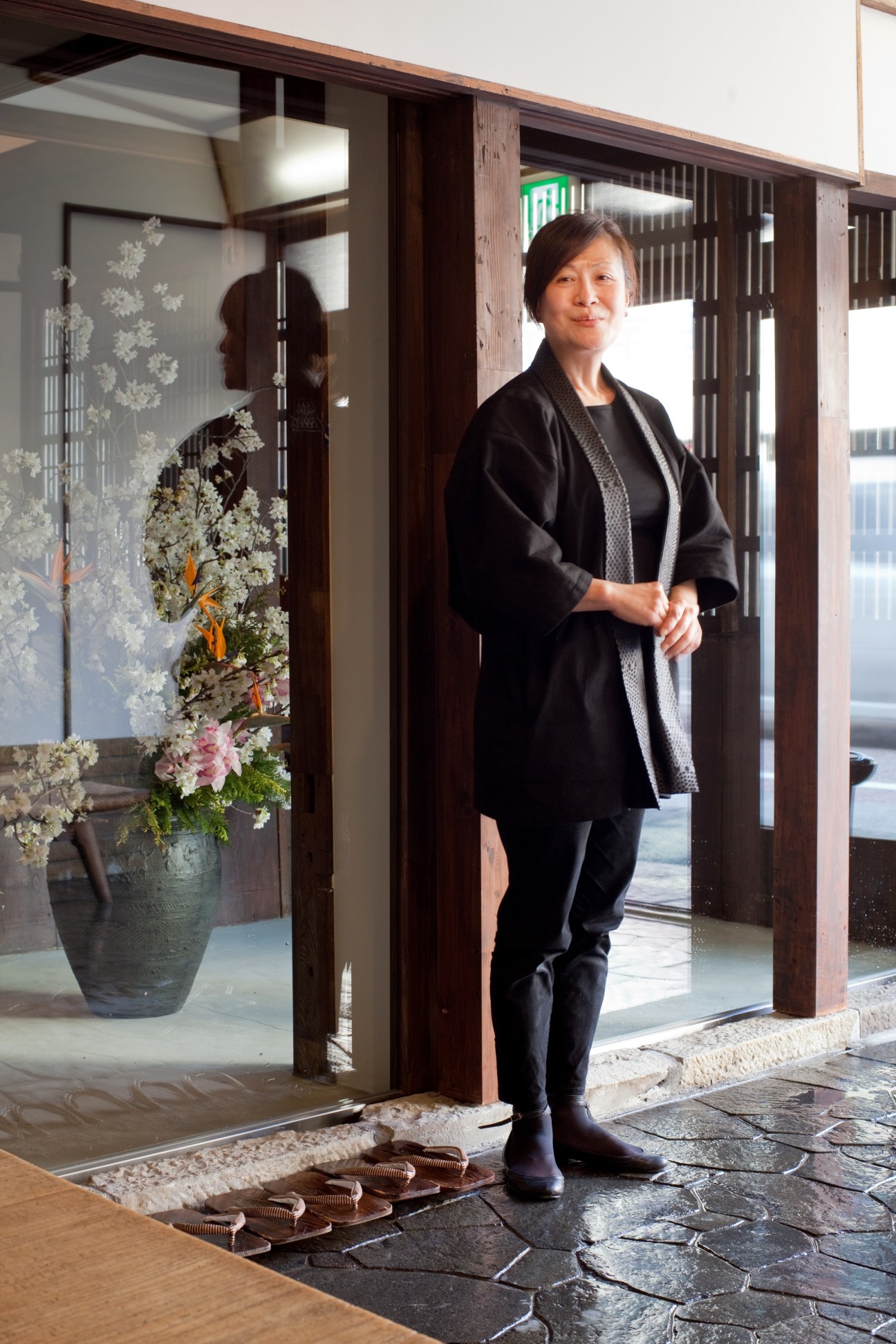 Mari Mori, Kiya Ryokan's chief service manager, stands in the renovated ryokan's entrance with wooden lattice doors and floral arrangement.