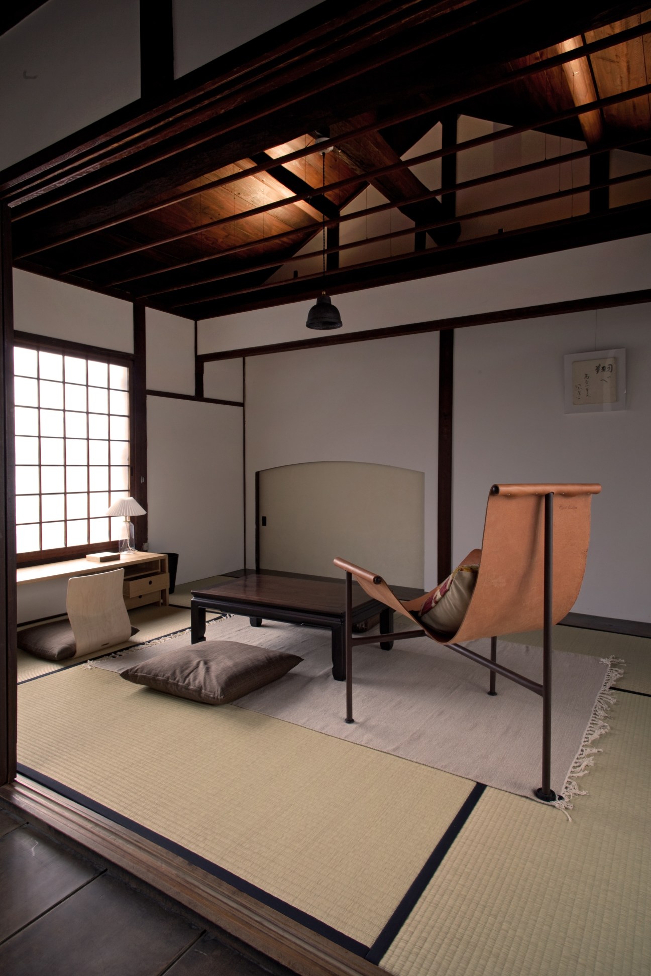 Interior of a traditional Japanese room with tatami mats, wooden beams, lattice window, low table, floor cushion, and modern leather chair.