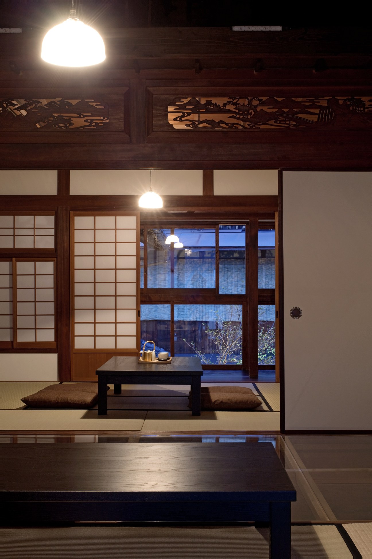 Interior of Kiya Ryokan showing traditional wooden screens, pendant lights, low table with cushions, and garden view at dusk.