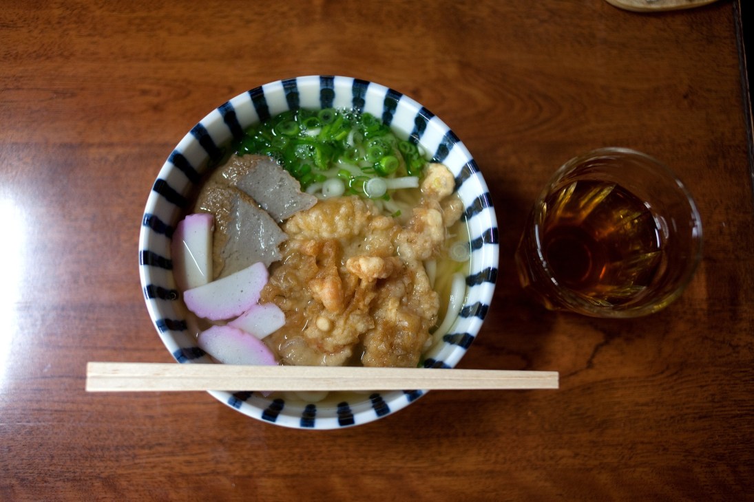 Bowl of udon noodles with broth, meat, and green onions on wooden table with chopsticks and drink.