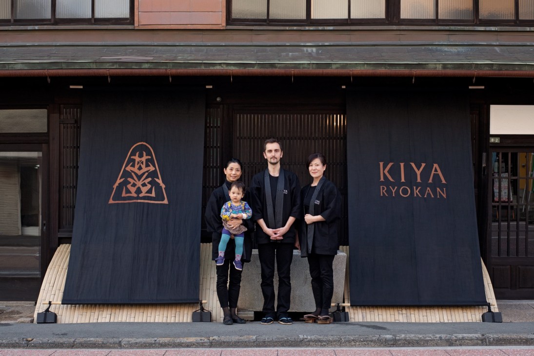 Four people stand in front of Kiya Ryokan's dark wooden facade with the ryokan's name and logo displayed on panels.