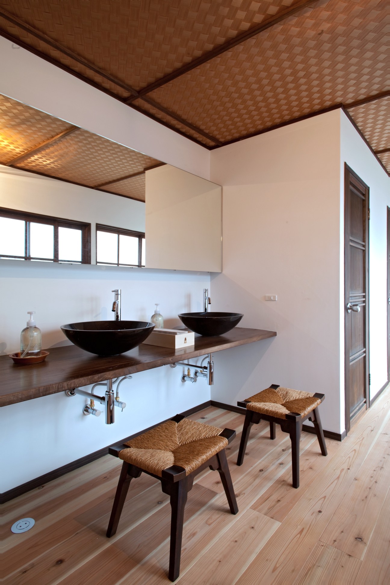 Modern bathroom with two black vessel sinks on wooden counter, woven ceiling panels, and woven rush-seat stools.