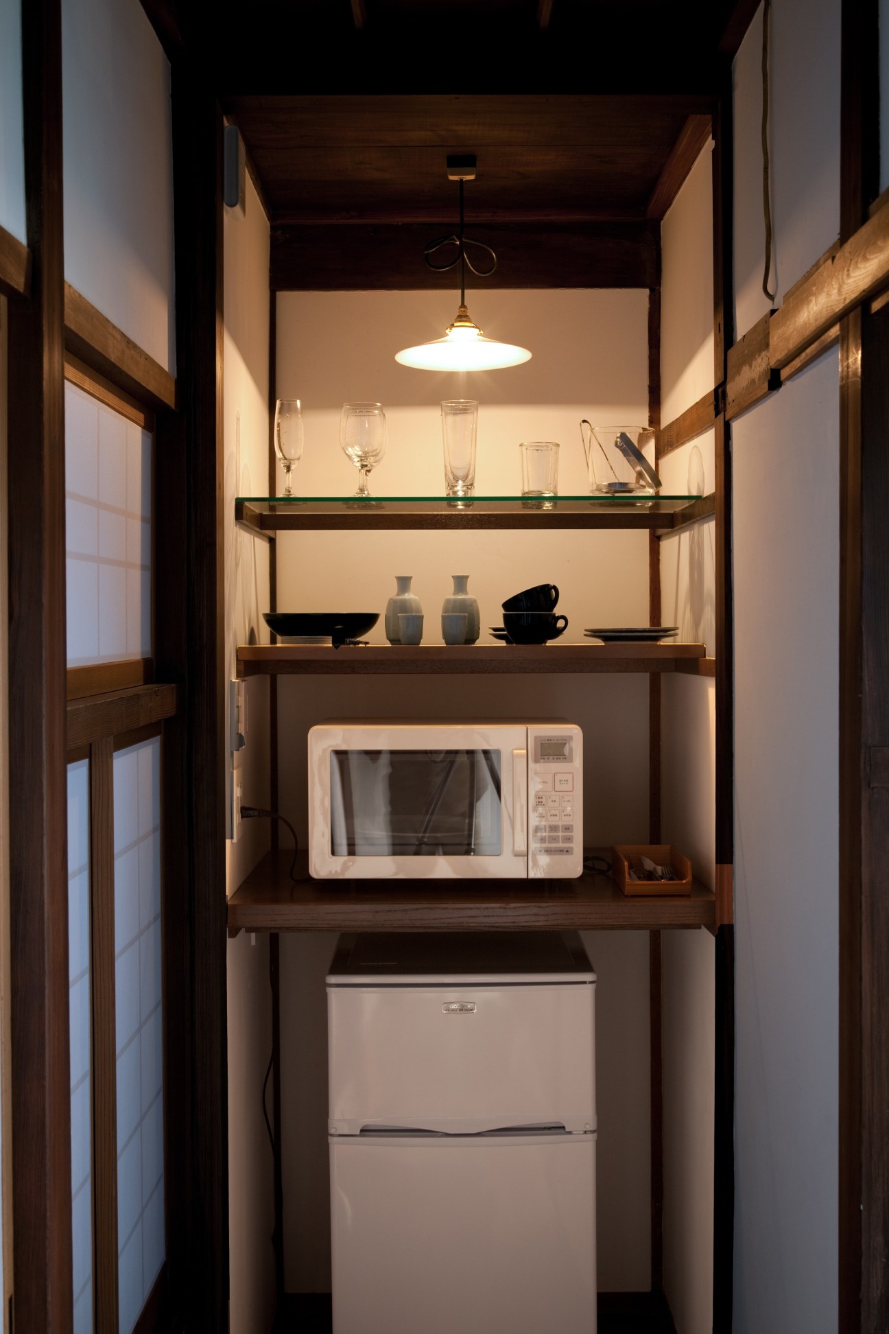A narrow communal storage closet with wooden shelving holding glassware, dishes, a microwave and refrigerator, illuminated by a vintage pendant light.