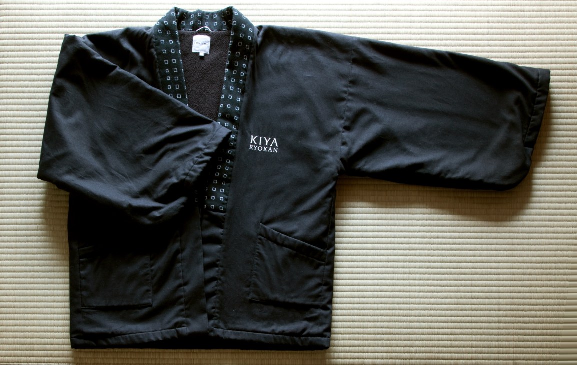 Black fleece-lined hanten jacket with Kiya Ryokan branding on tatami mat background.