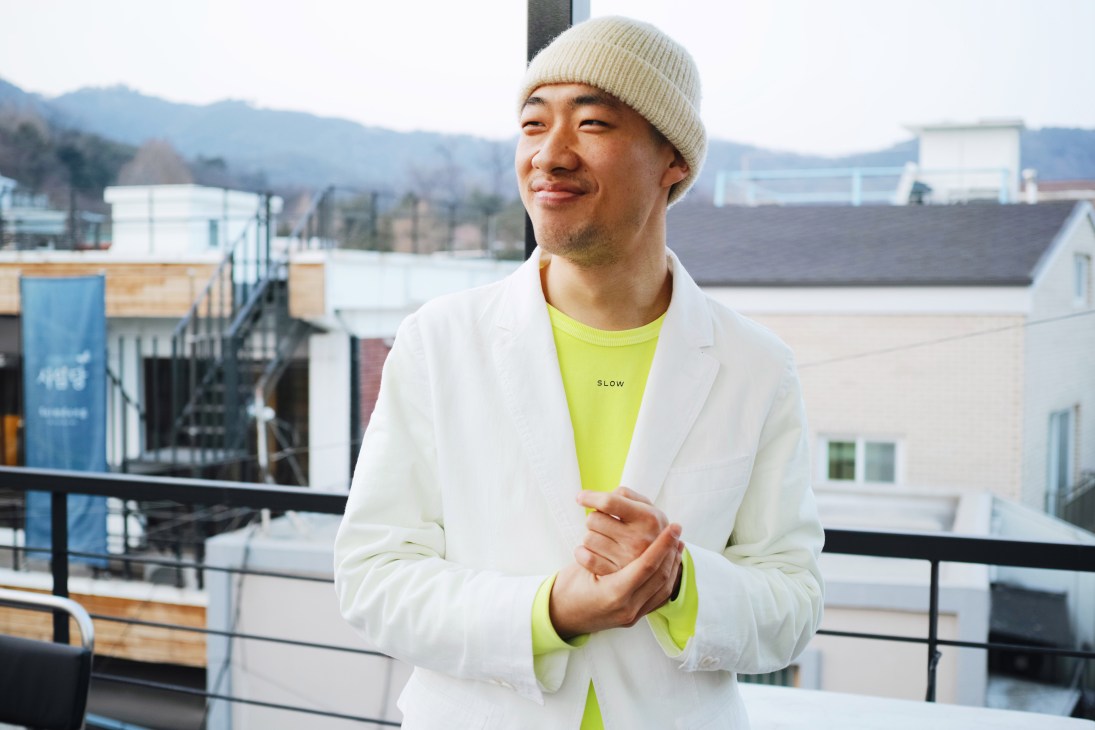 Man in cream beanie and white blazer over neon yellow t-shirt on rooftop terrace with Seoul buildings and mountains behind.