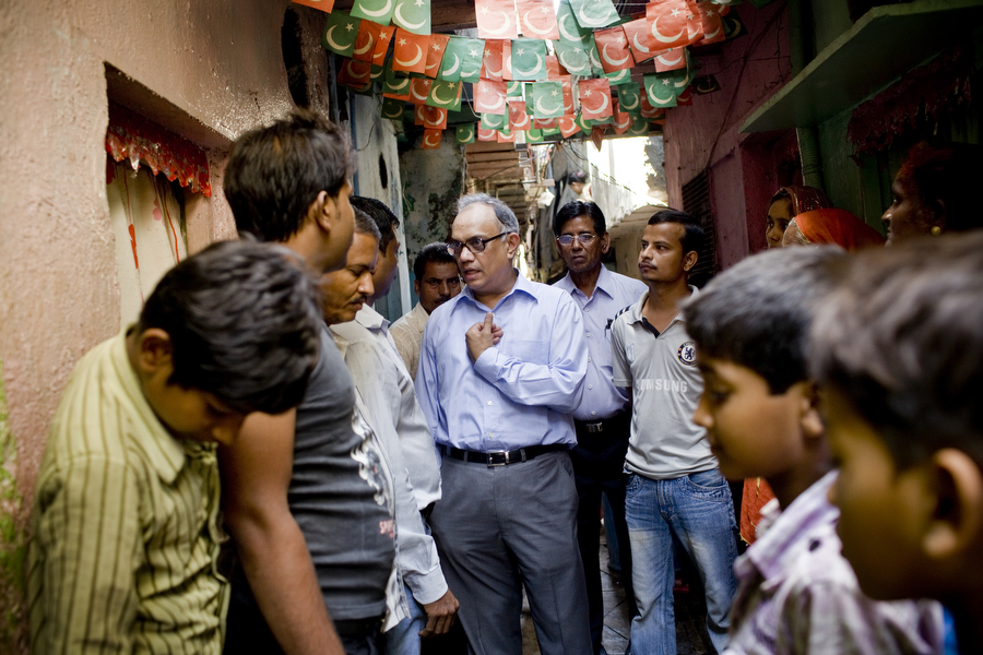 Mukesh Mehta in a light blue shirt speaks with residents in a narrow Mumbai alleyway decorated with colorful bunting.