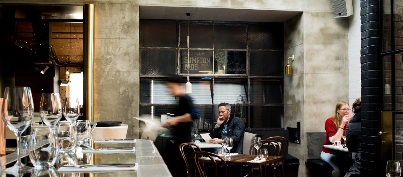 Interior of Smalls Bar Melbourne showing diners at tables with wine glasses and a server passing by a large window.