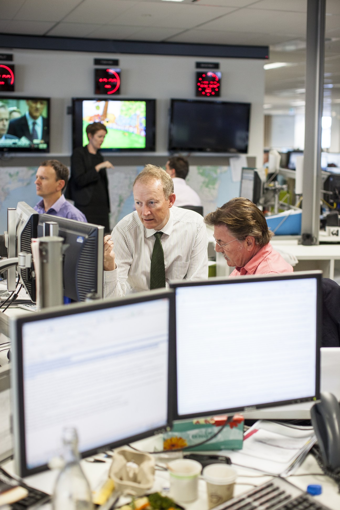 A man in a white shirt and green tie sits at a desk in a newsroom with computer monitors and colleagues visible in the background.