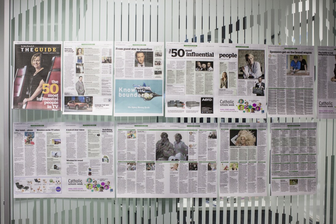 Newspaper pages displayed on a vertical ribbed wall showing Sydney Morning Herald layouts and sections.