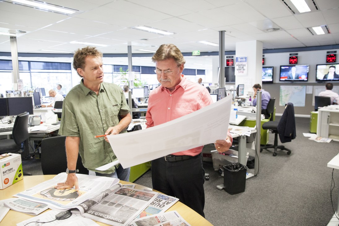 Two men in a newsroom examine a newspaper layout together at a desk.