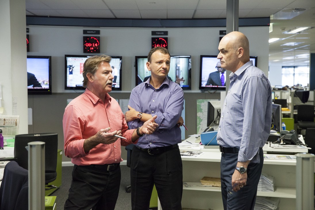 Three men in business casual attire converse in a modern newsroom with multiple monitors displaying news feeds in the background.