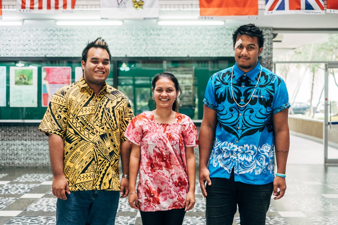 Three international students in colorful patterned shirts stand in a university corridor with international flags hanging above.