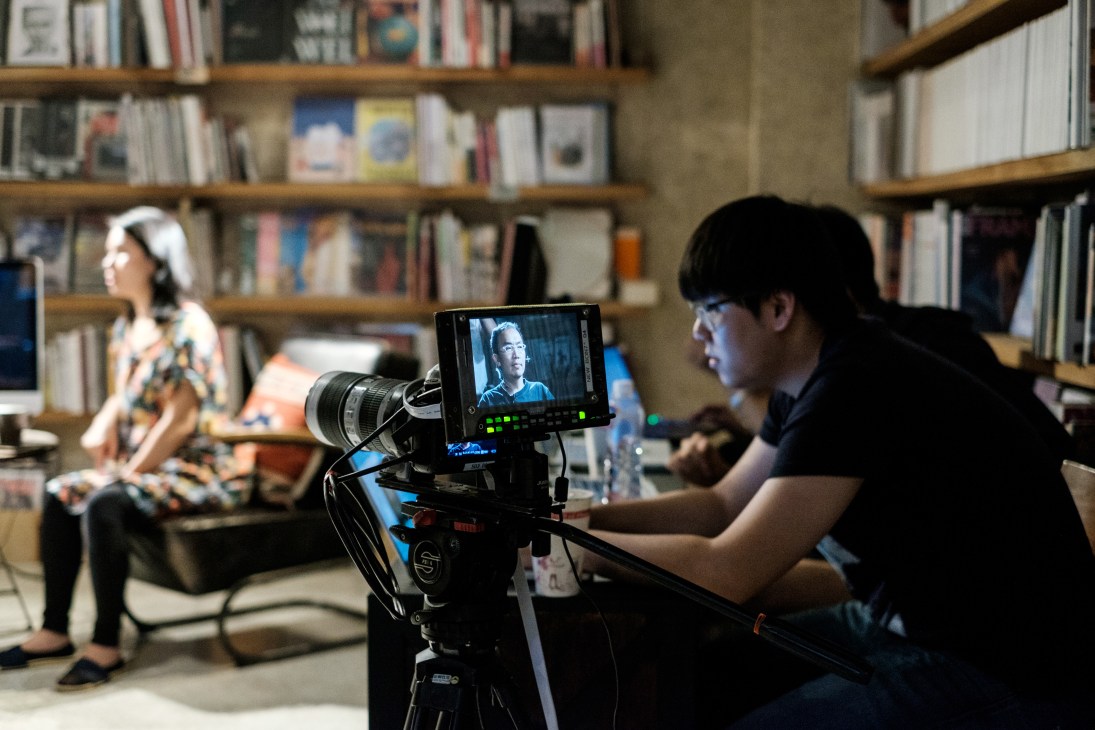 Cameraman operating television camera in library studio with bookshelves and woman in background.