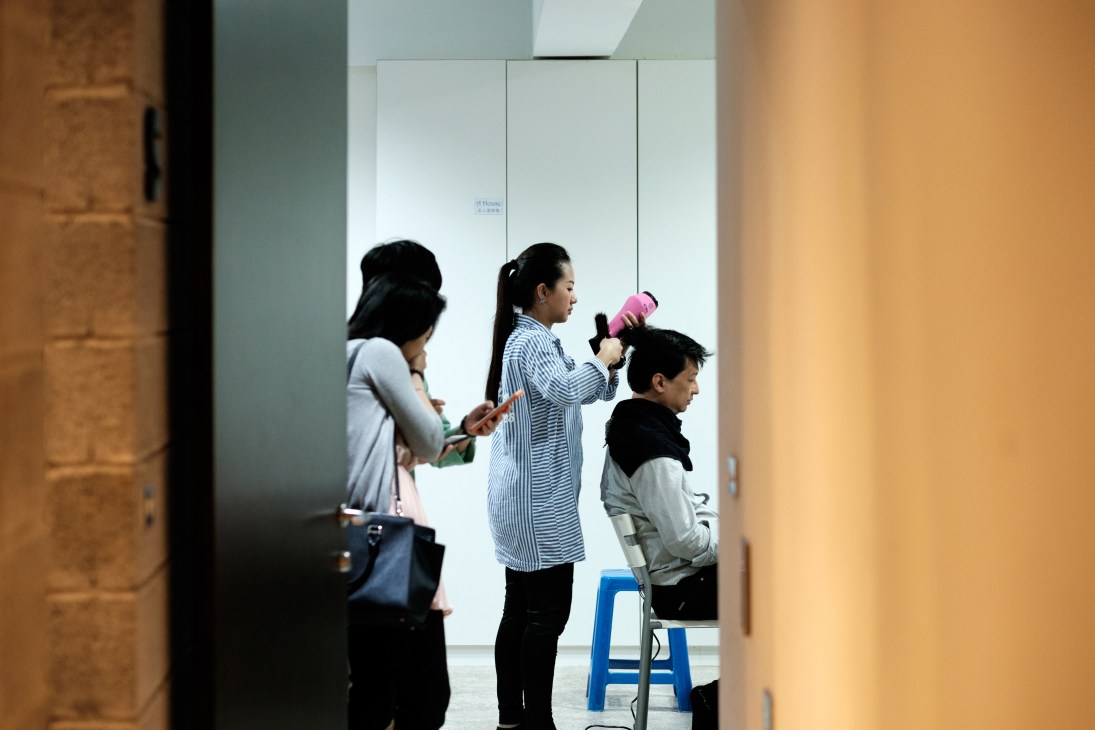 A woman with a blow dryer styles a man's hair while another woman watches in a bright studio space.