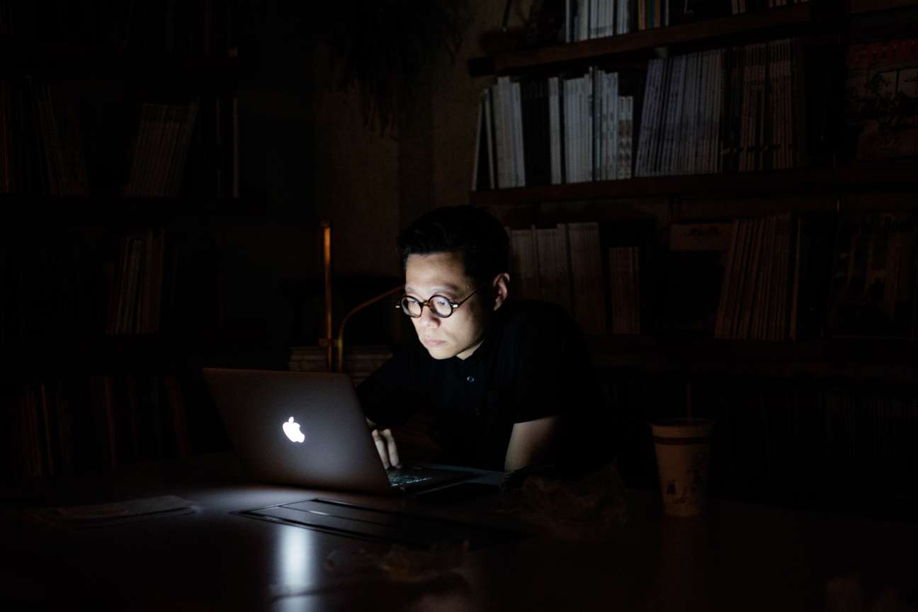 Art director Yu Feng wearing glasses works on a laptop in a dark library setting with bookshelves behind.