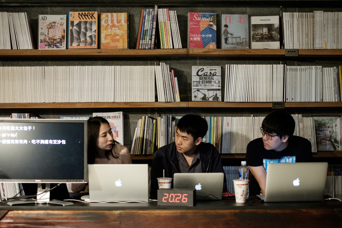 Three people work at laptops in front of shelves of magazines and design publications in a library setting.
