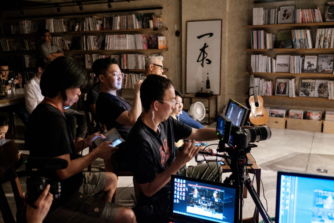 Production crew setting up camera equipment and monitoring screens in a magazine library studio with bookshelves.
