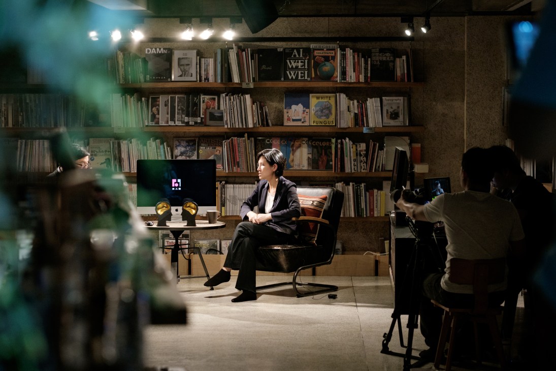 Woman in dark suit seated on black chair in studio with bookshelves behind, being interviewed on camera.