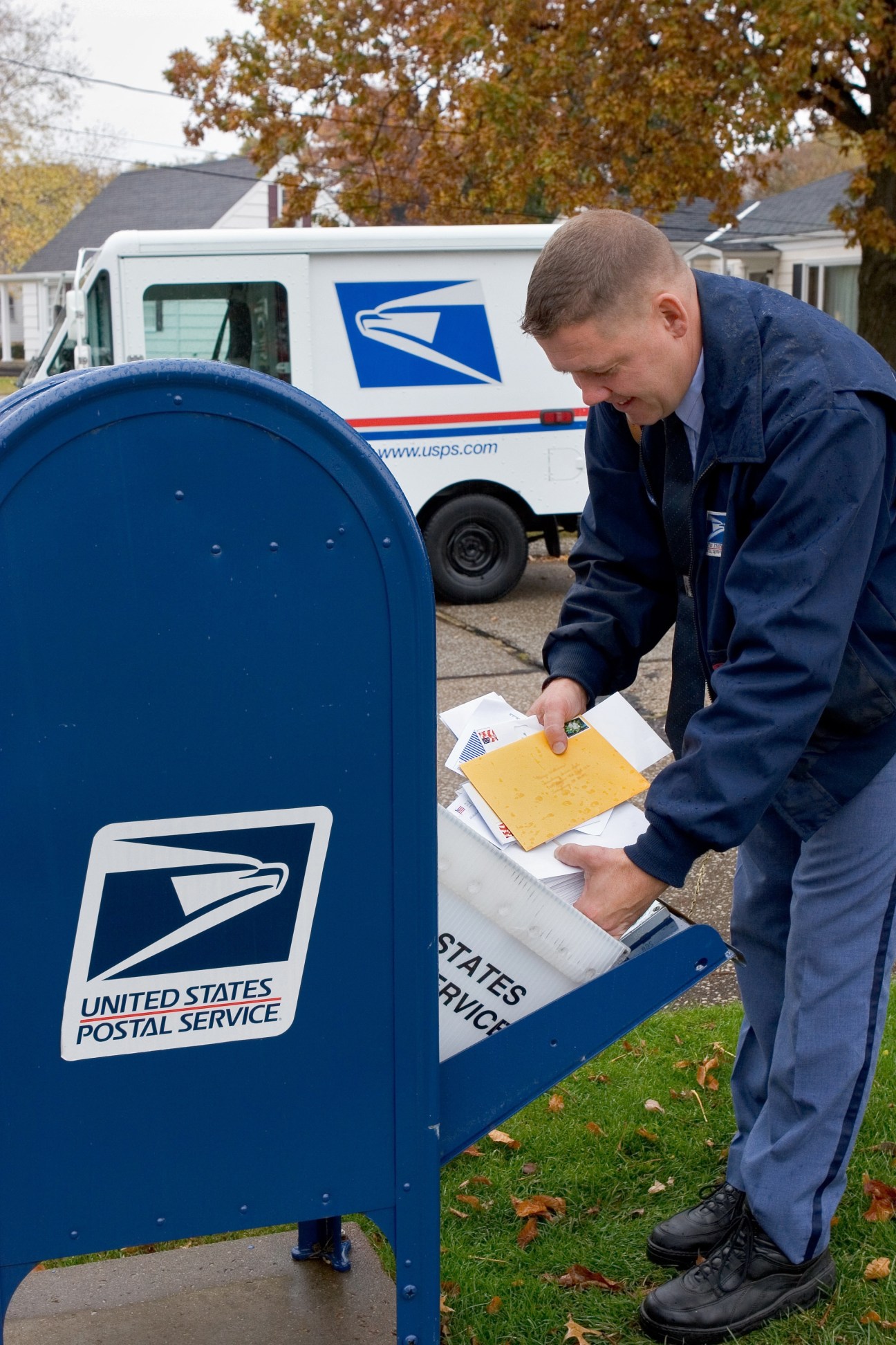 US Postal Service worker depositing mail into a blue USPS mailbox with delivery truck in background.