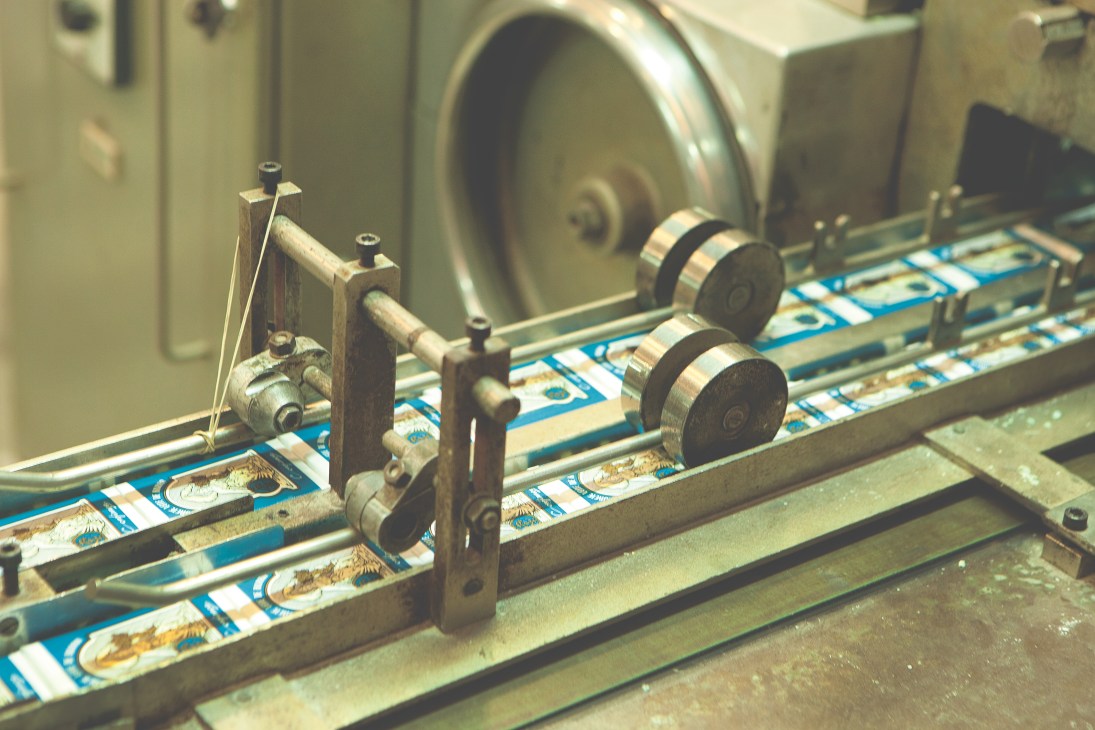 Vintage mechanical sorting and labelling machine with metal rollers guiding packaged Claus Porto soap bars along a conveyor belt.