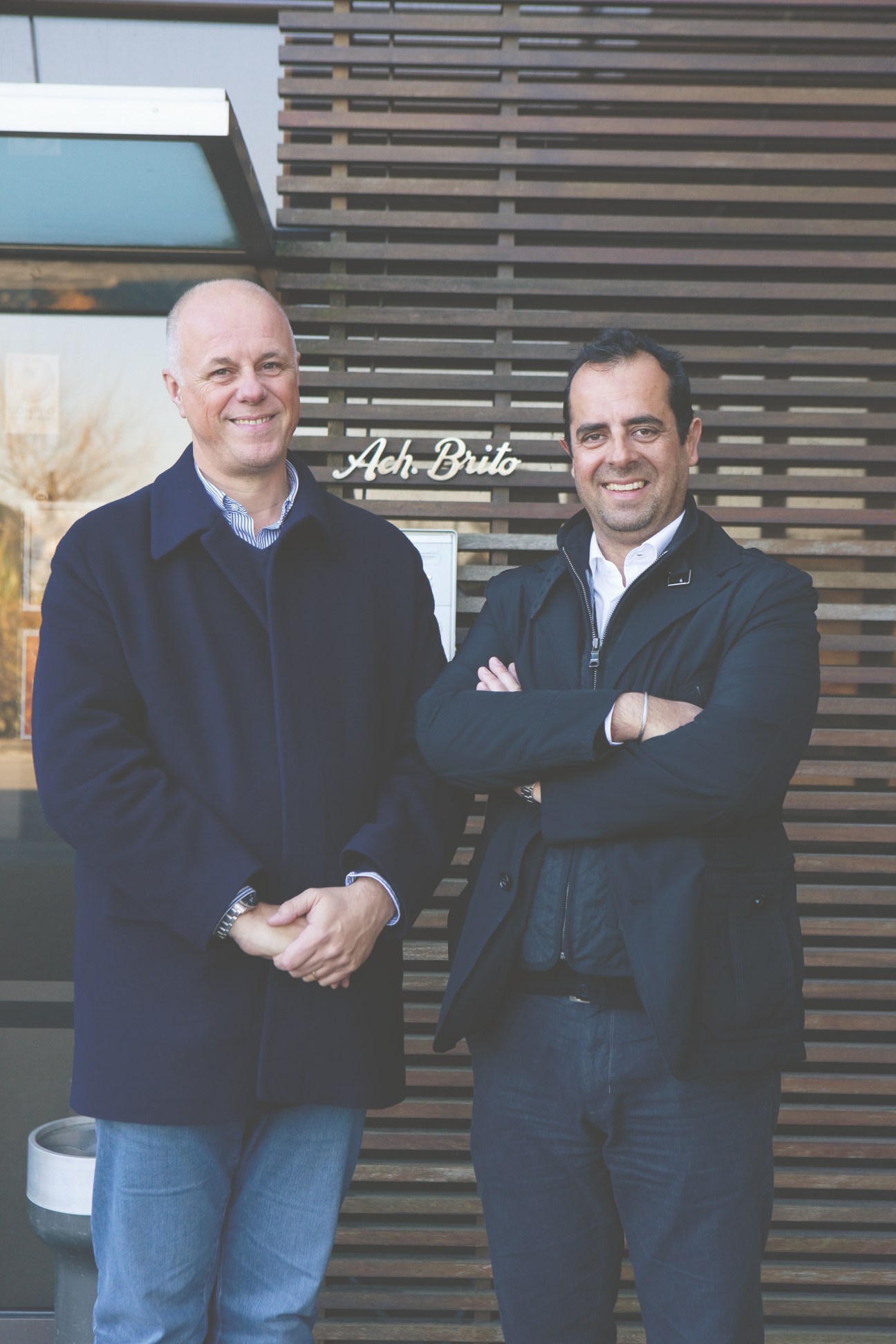 José Fernandes and Aquiles Brito stand together smiling in front of the Ach. Brito factory entrance.