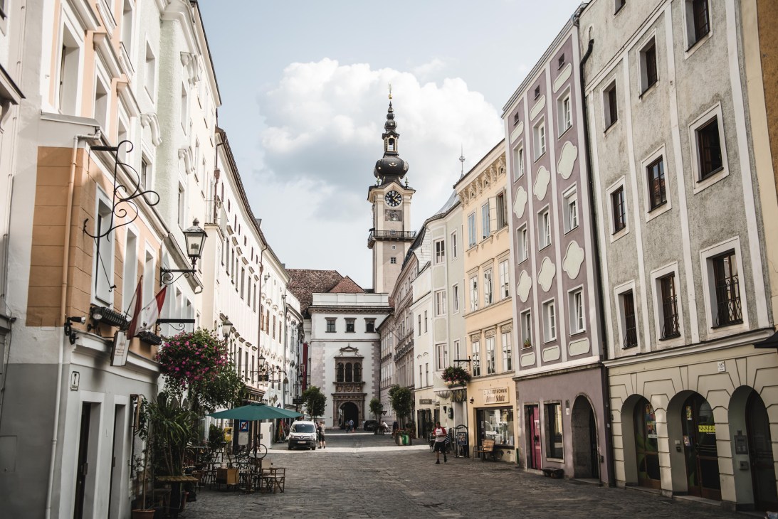 Historic cobblestone square in Linz with colorful baroque buildings, outdoor cafe seating, and clock tower
