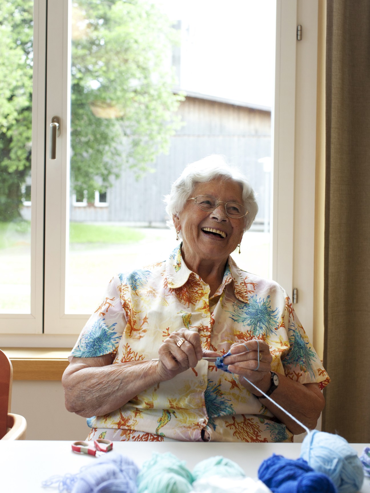 Elderly woman smiling while knitting by a large window with countryside visible behind her.