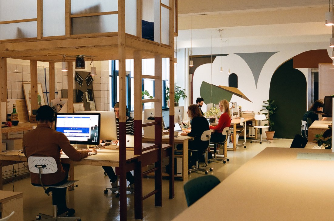 Modern open office workspace with wooden structural beams, employees at desks, and green sculptural wall elements.