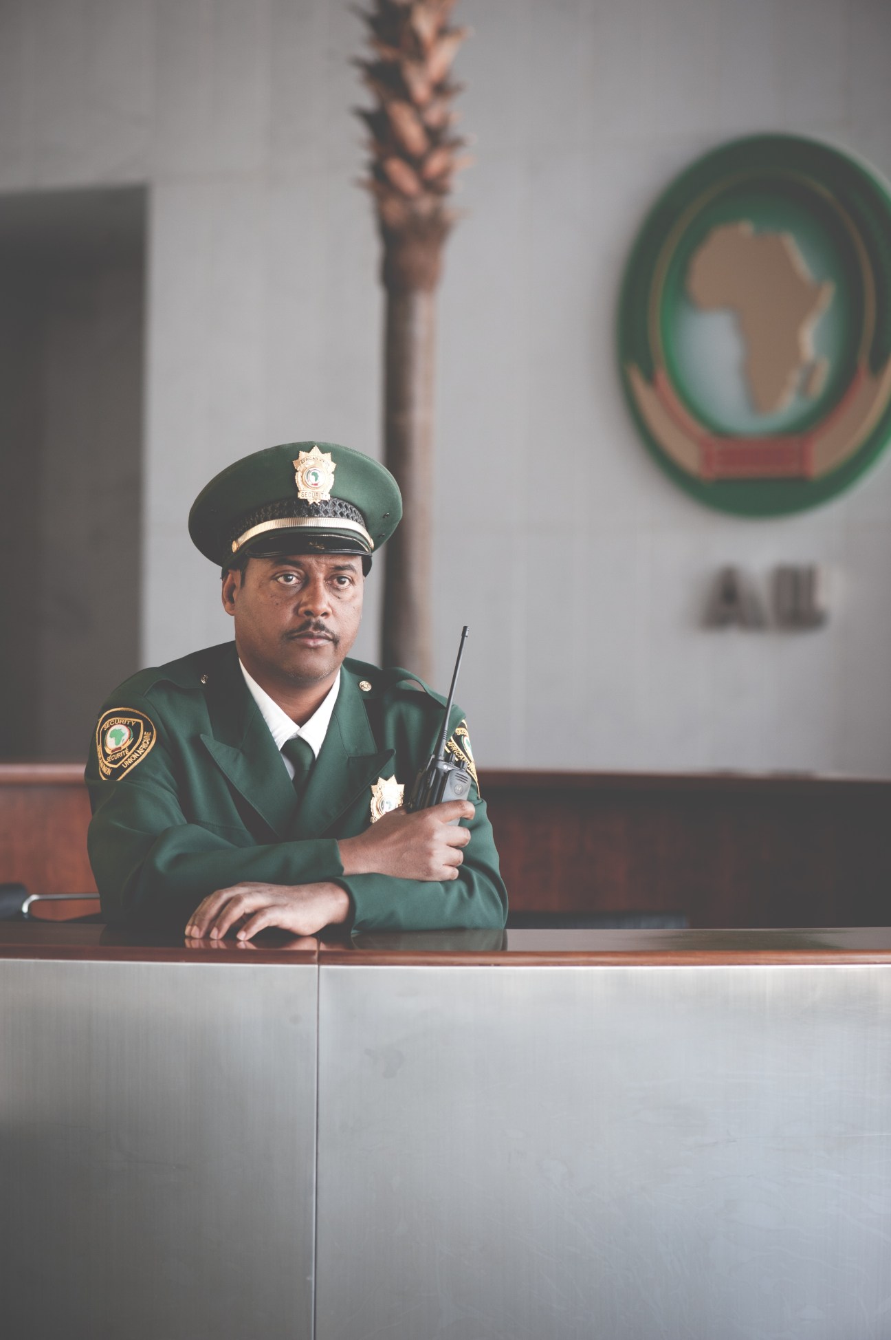 Security guard in green uniform with cap and radio standing at African Union entrance desk.