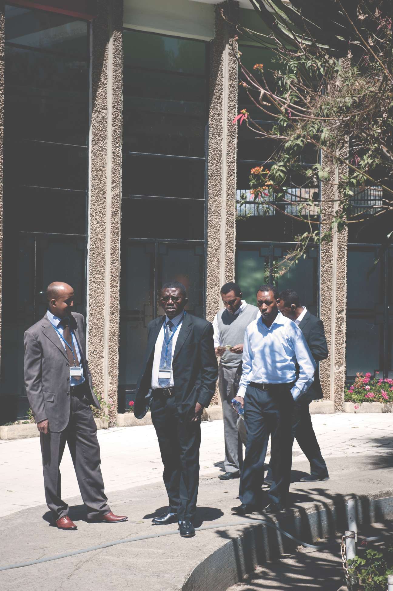 Five men in business suits walk outside a modern building with large windows and stone columns.