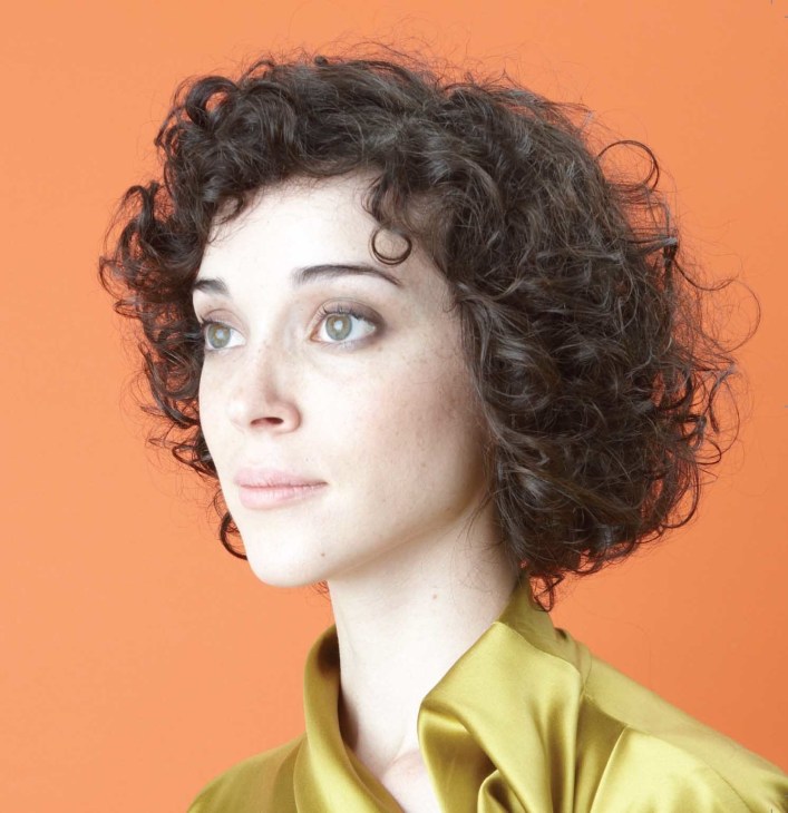 St Vincent, a woman with curly dark hair and pale complexion wearing a yellow-green garment against an orange background.