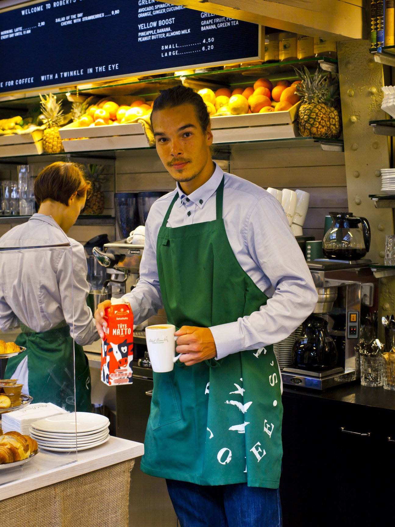 Barista in green apron holding a cup at Robert's coffee counter with fresh produce displayed behind.