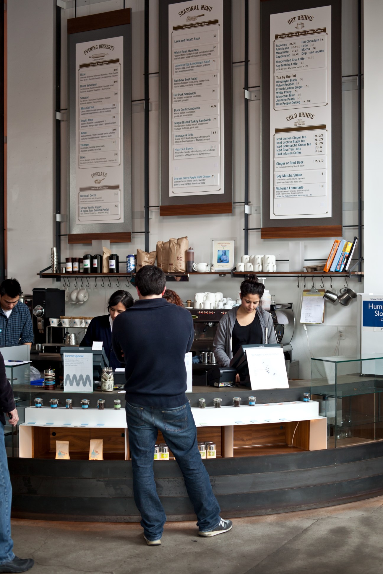 Customer ordering at a modern coffee shop counter with menu boards displayed on the wall above.