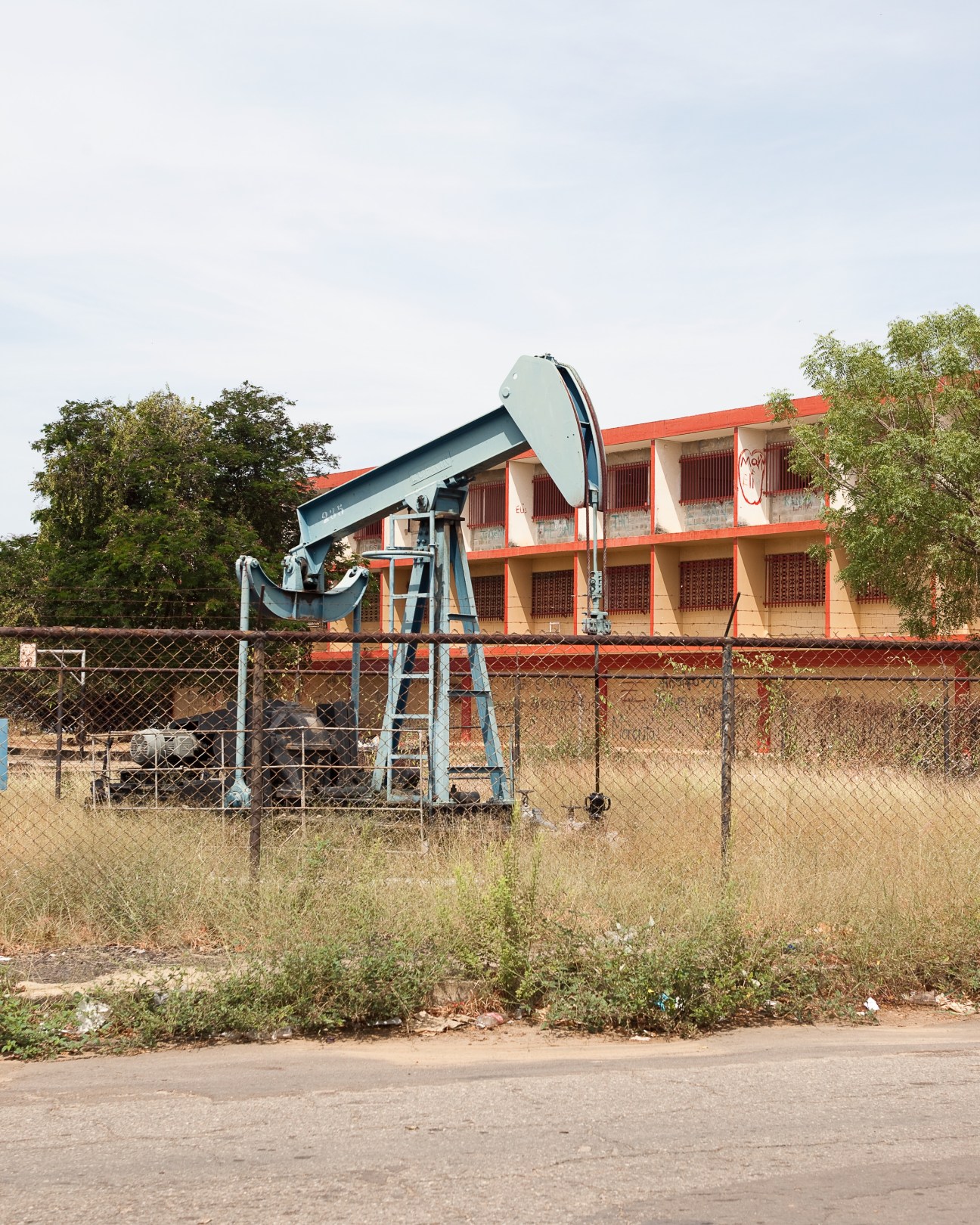 Oil pumpjack extracting crude from the ground in Cabimas, Venezuela, with an orange and white building behind a chain-link fence.