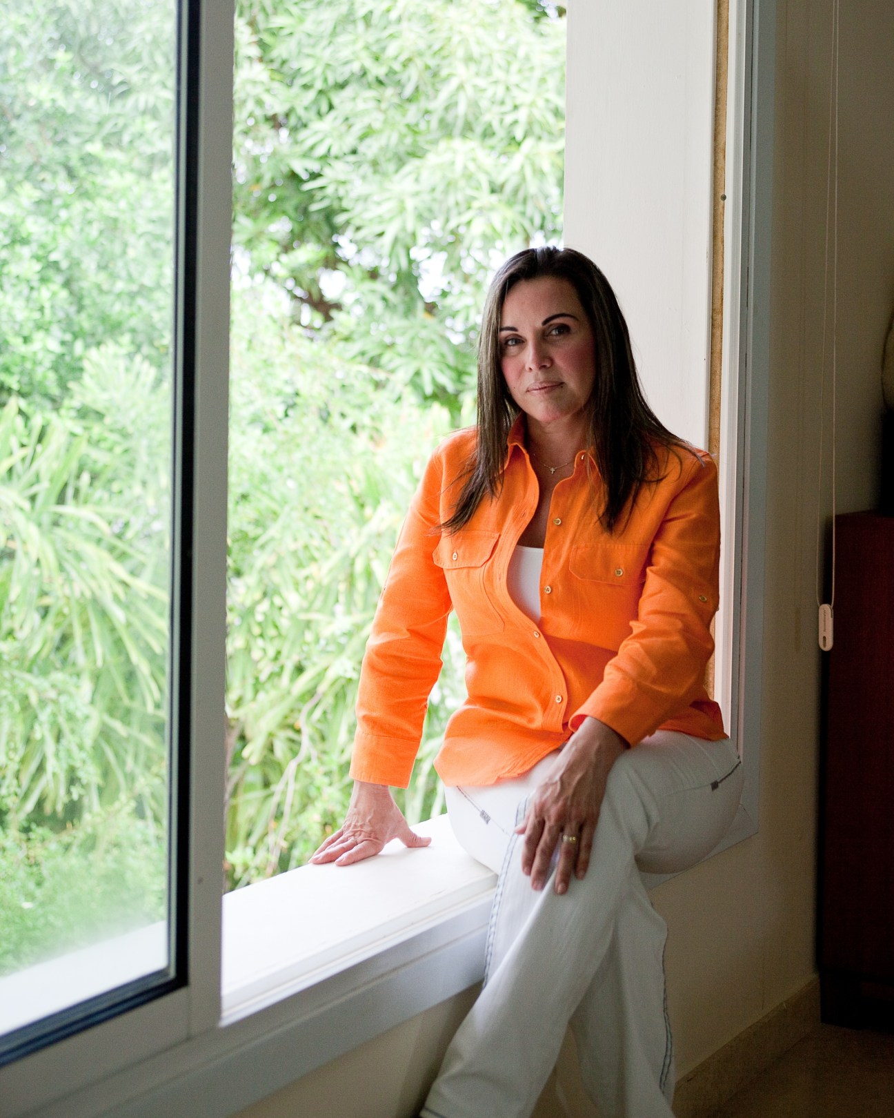 Woman in orange shirt sitting by window with green foliage visible outside