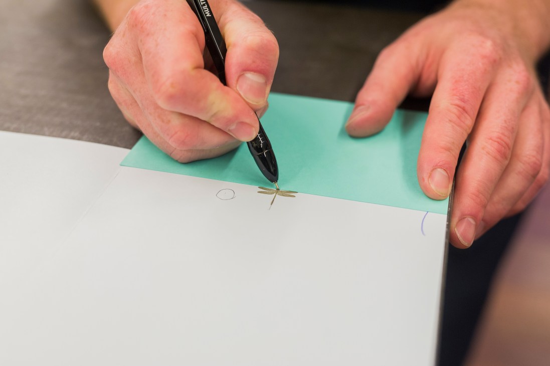 A person uses a thin tool to adjust printed details on white and turquoise paper sheets.