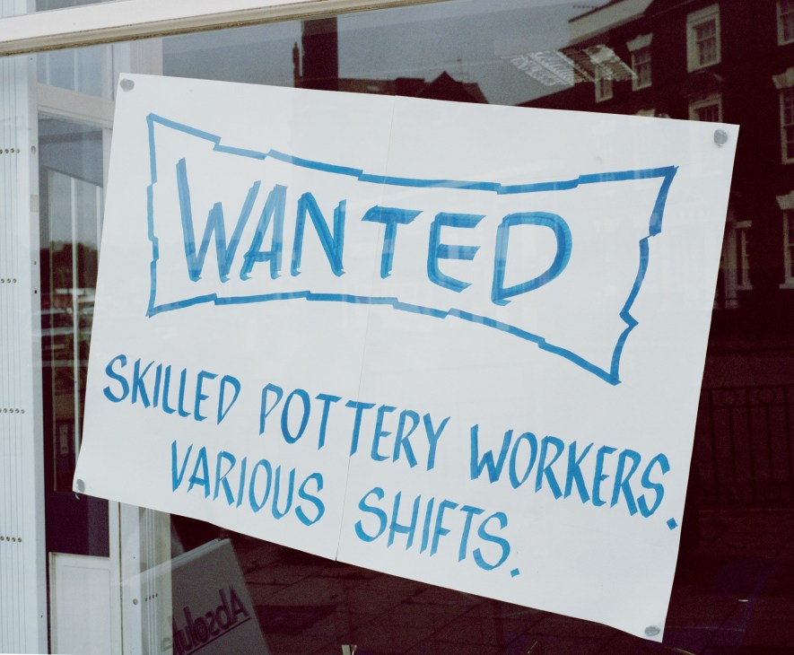 A handwritten sign in a window reading 'WANTED: SKILLED POTTERY WORKERS VARIOUS SHIFTS' in blue marker on white paper.