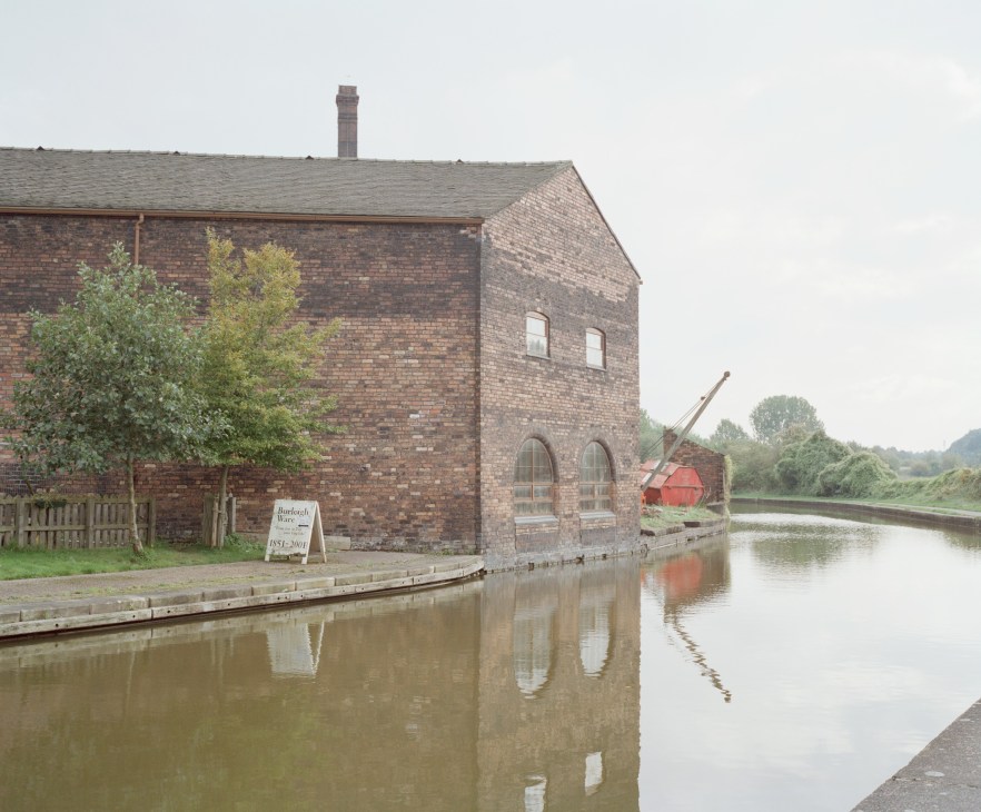 Historic brick pottery building reflected in still canal water with red industrial equipment visible.