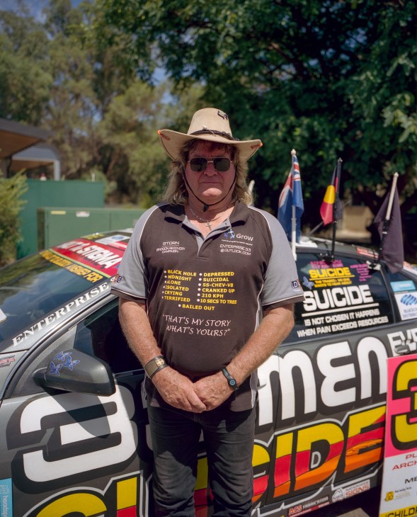 Storn Petterson stands in front of his decorated vehicle wearing a wide-brimmed hat and sunglasses, displaying a message shirt about rural isolation.