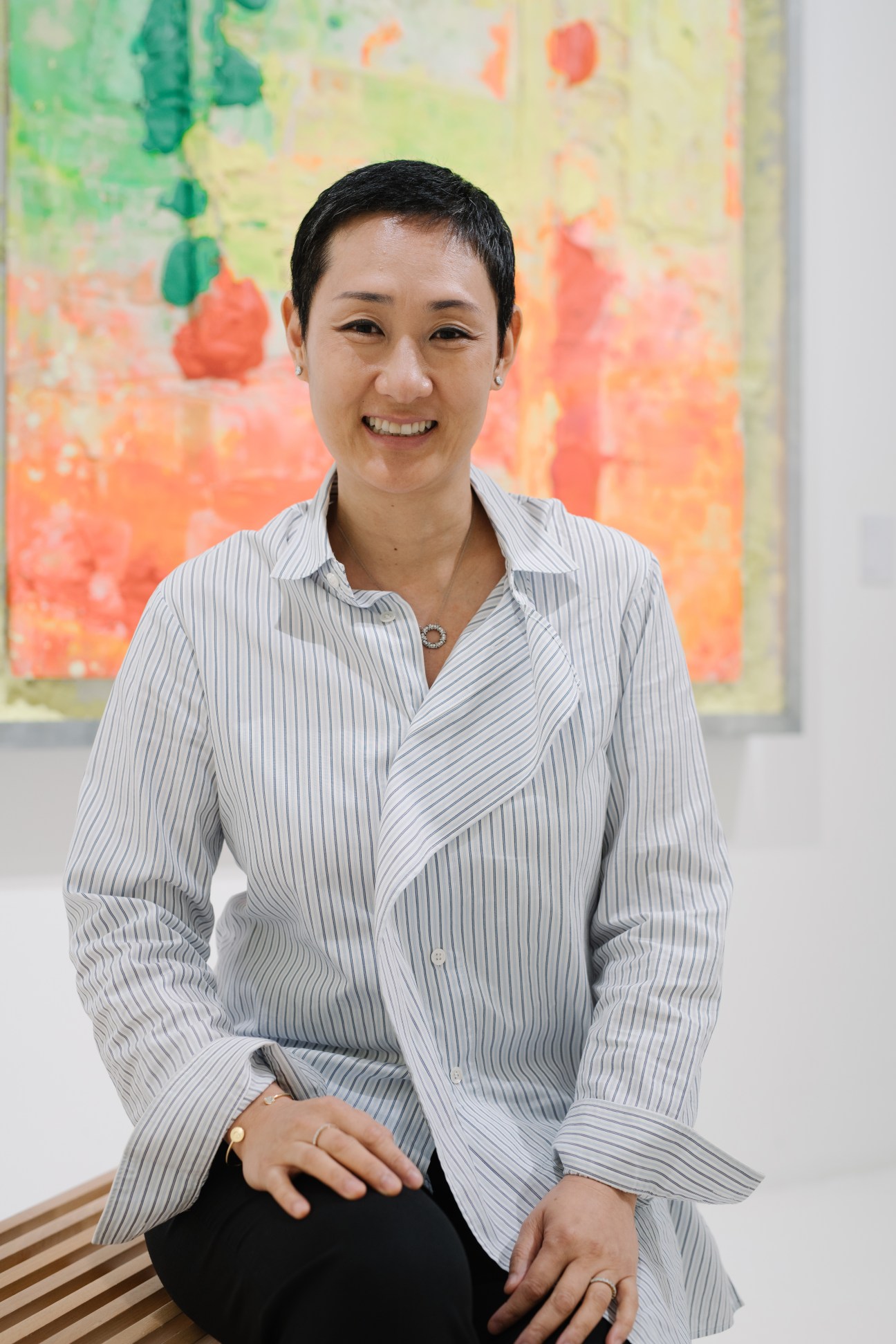 Emi Eu, STPI director, smiling in front of colorful abstract artwork in a gallery setting.