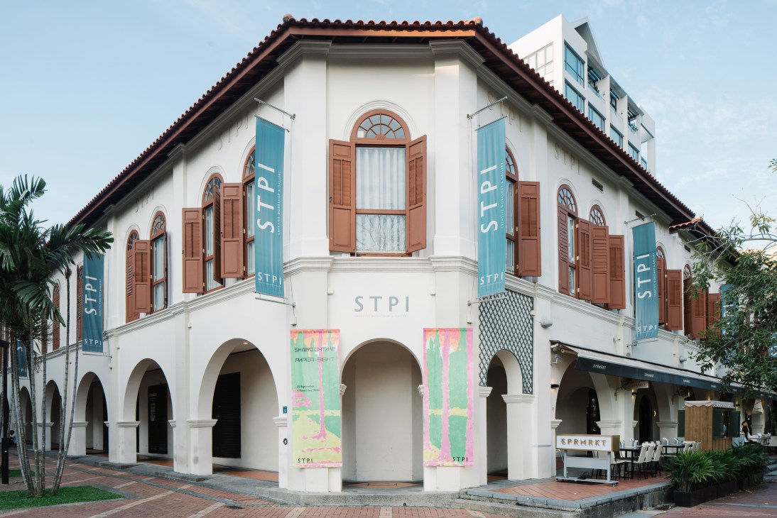 White colonial-style building with red tile roof and brown wooden shutters, displaying STPI banners and colorful exhibition posters.