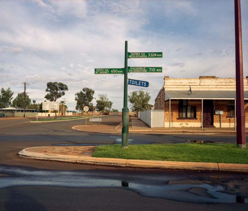 Directional sign in Cue, Australia showing distances to Darwin, Perth, and Kalgoorlie with a toilets sign nearby.