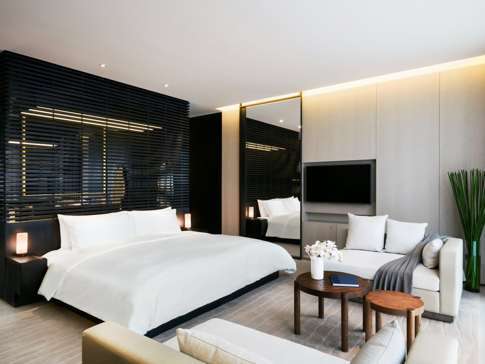 Modern hotel suite with white bed, cream sofa, black slatted wall panels with gold accents, wooden coffee table and green plant.