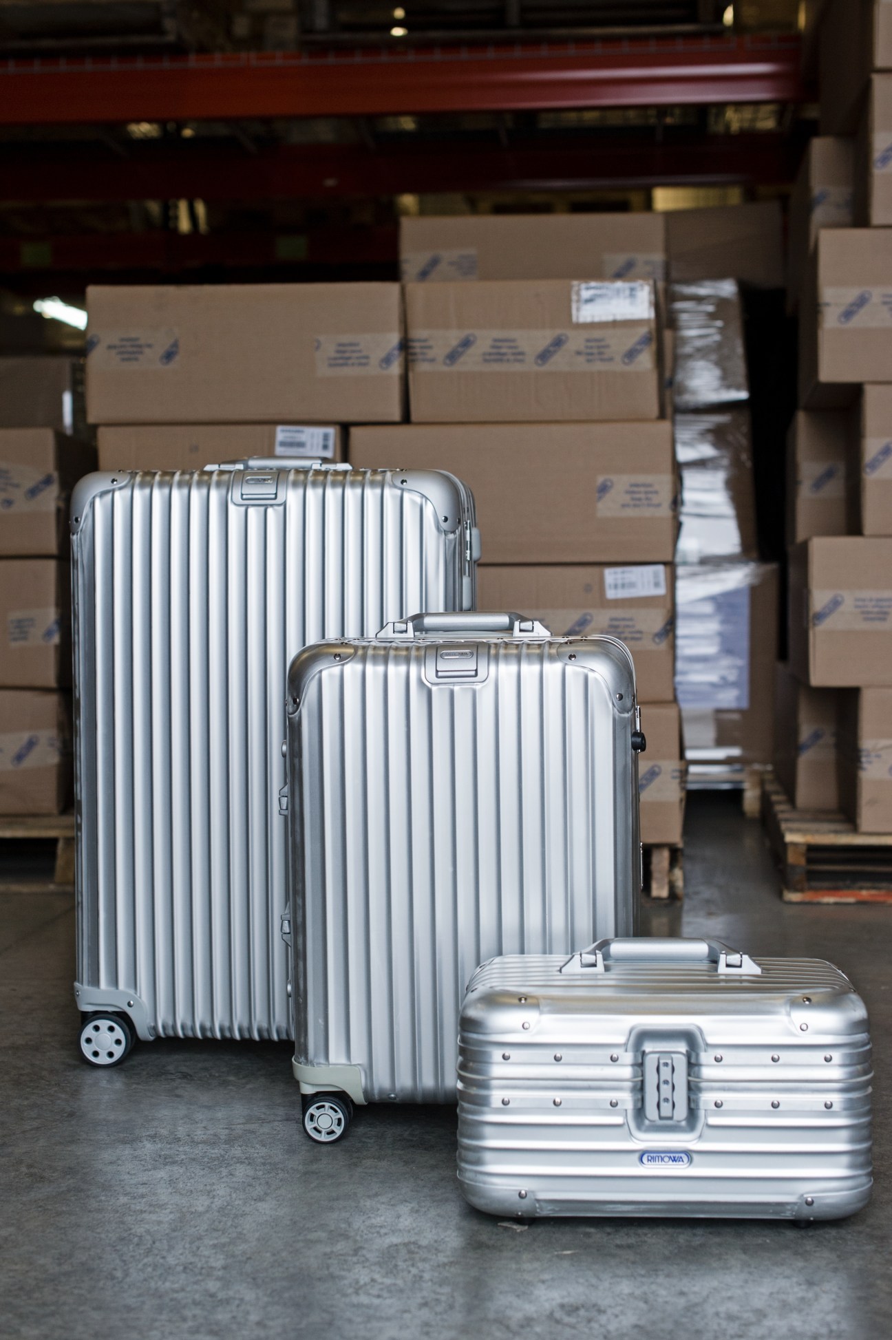 Three silver Rimowa aluminium suitcases of different sizes displayed in front of cardboard shipping boxes.