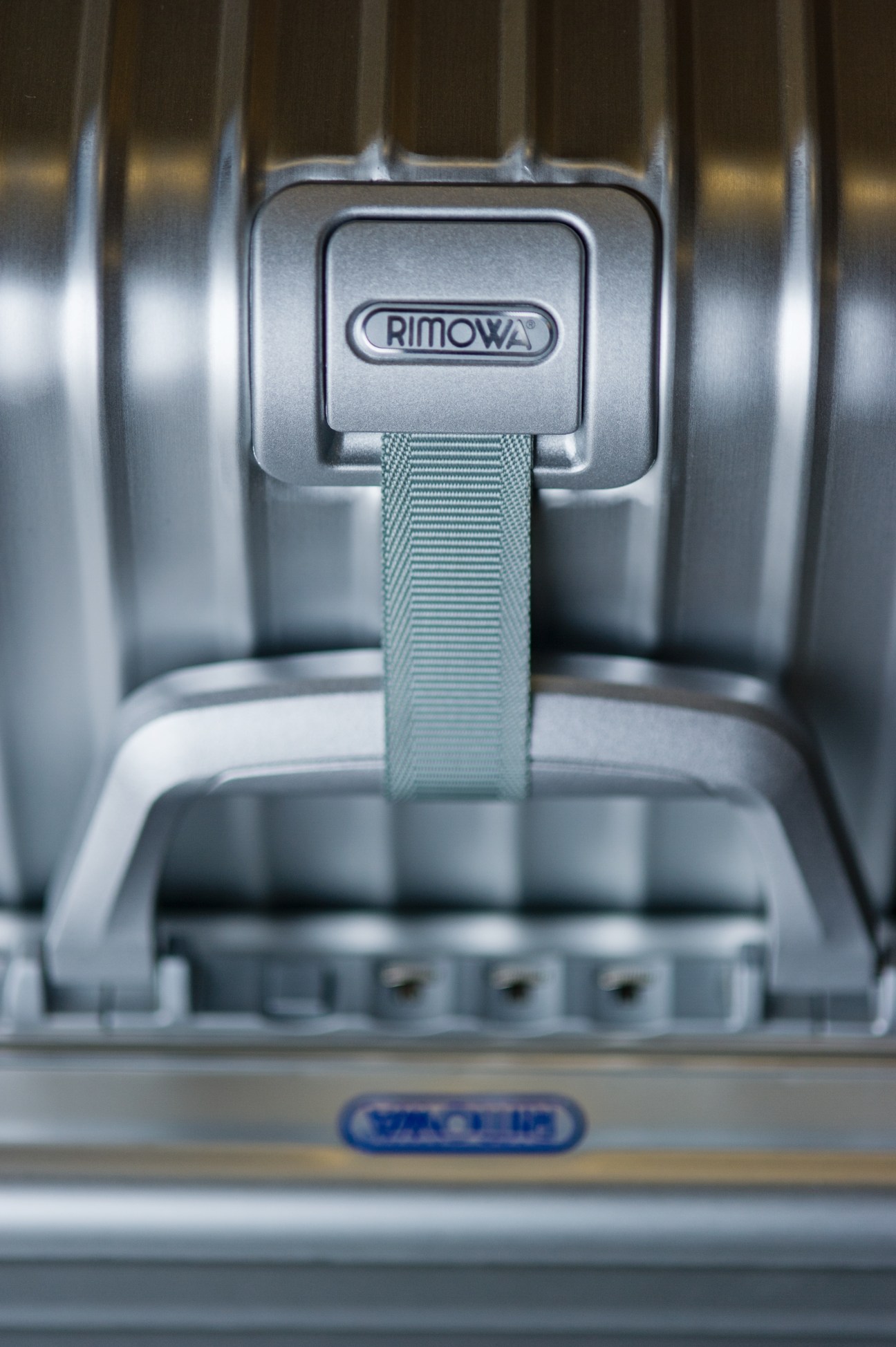 Close-up of a Rimowa suitcase handle with branded metal attachment and fabric strap.