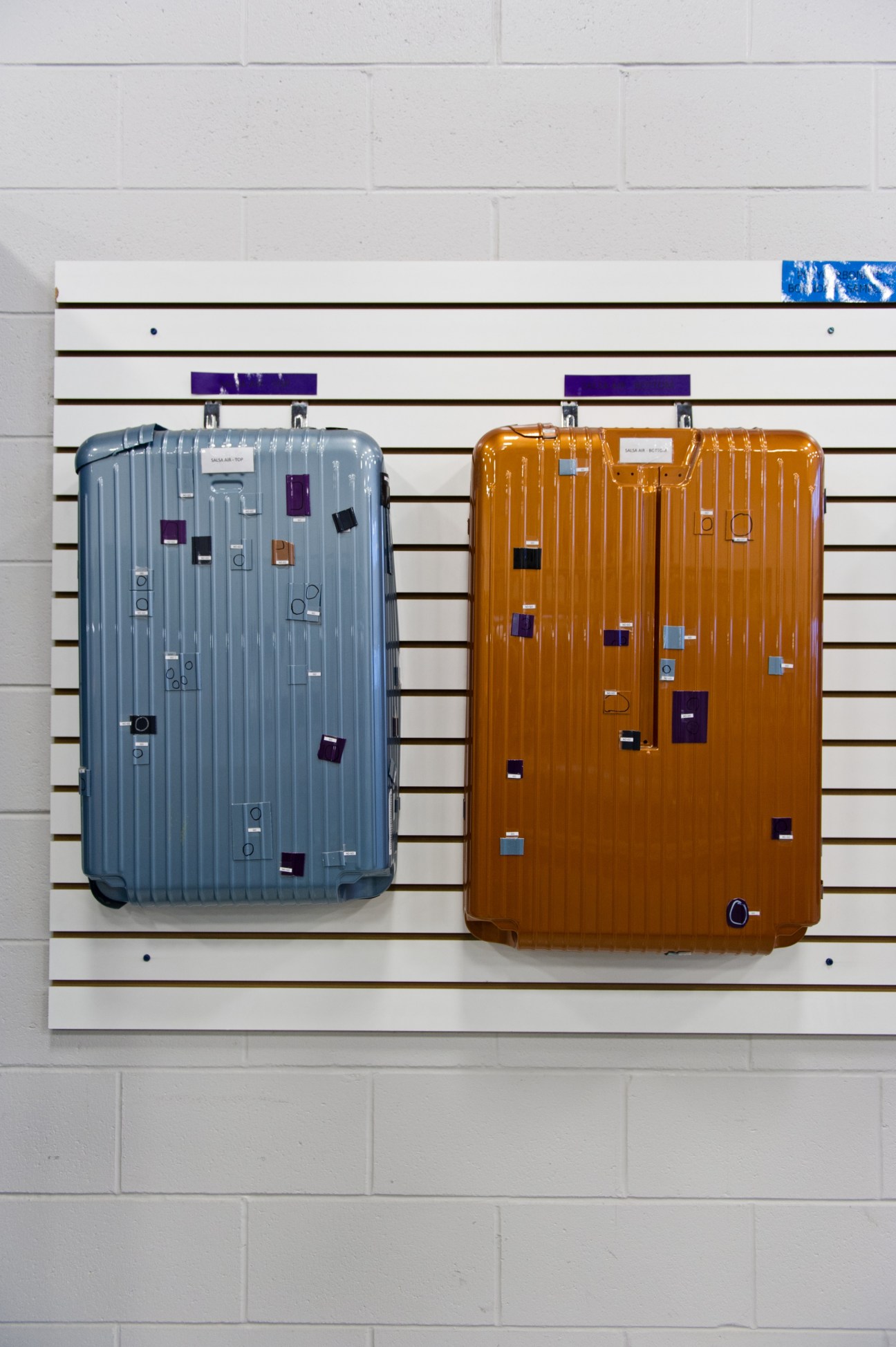 Two Rimowa hard-sided suitcases in blue and orange displayed on a white wall-mounted rack.