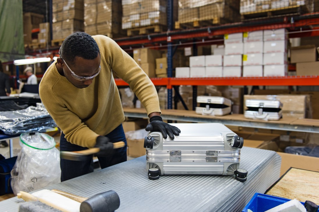 Worker in yellow sweater inspecting aluminum Rimowa suitcase in factory warehouse.